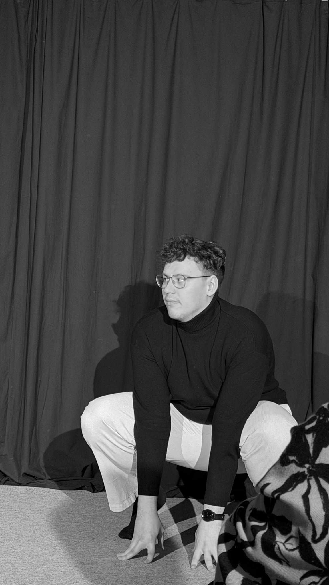A performer crouching alone in front of a stage curtain, in black and white